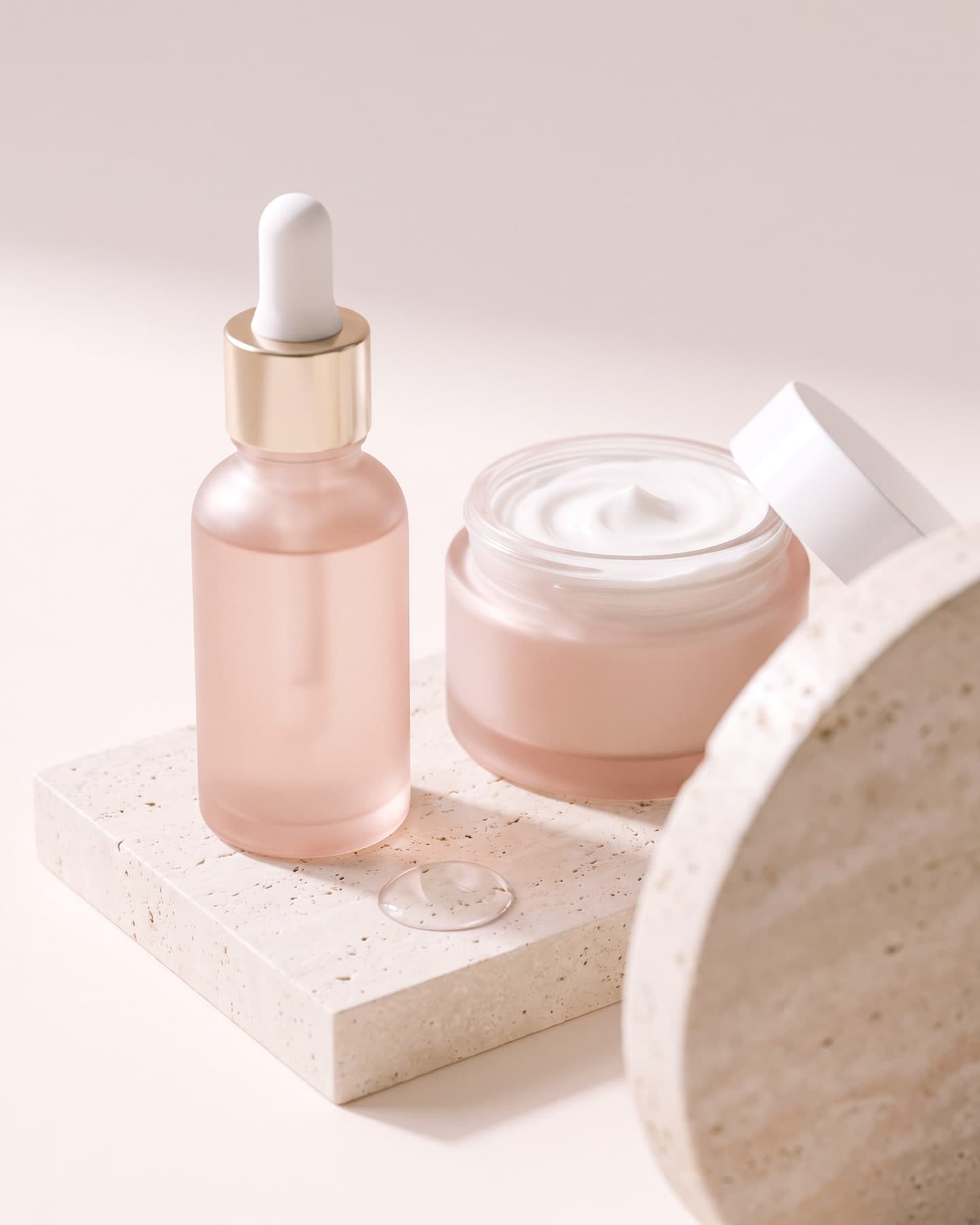 Frasco de sérum com conta-gotas e pote de creme aberto sobre pedra travertino